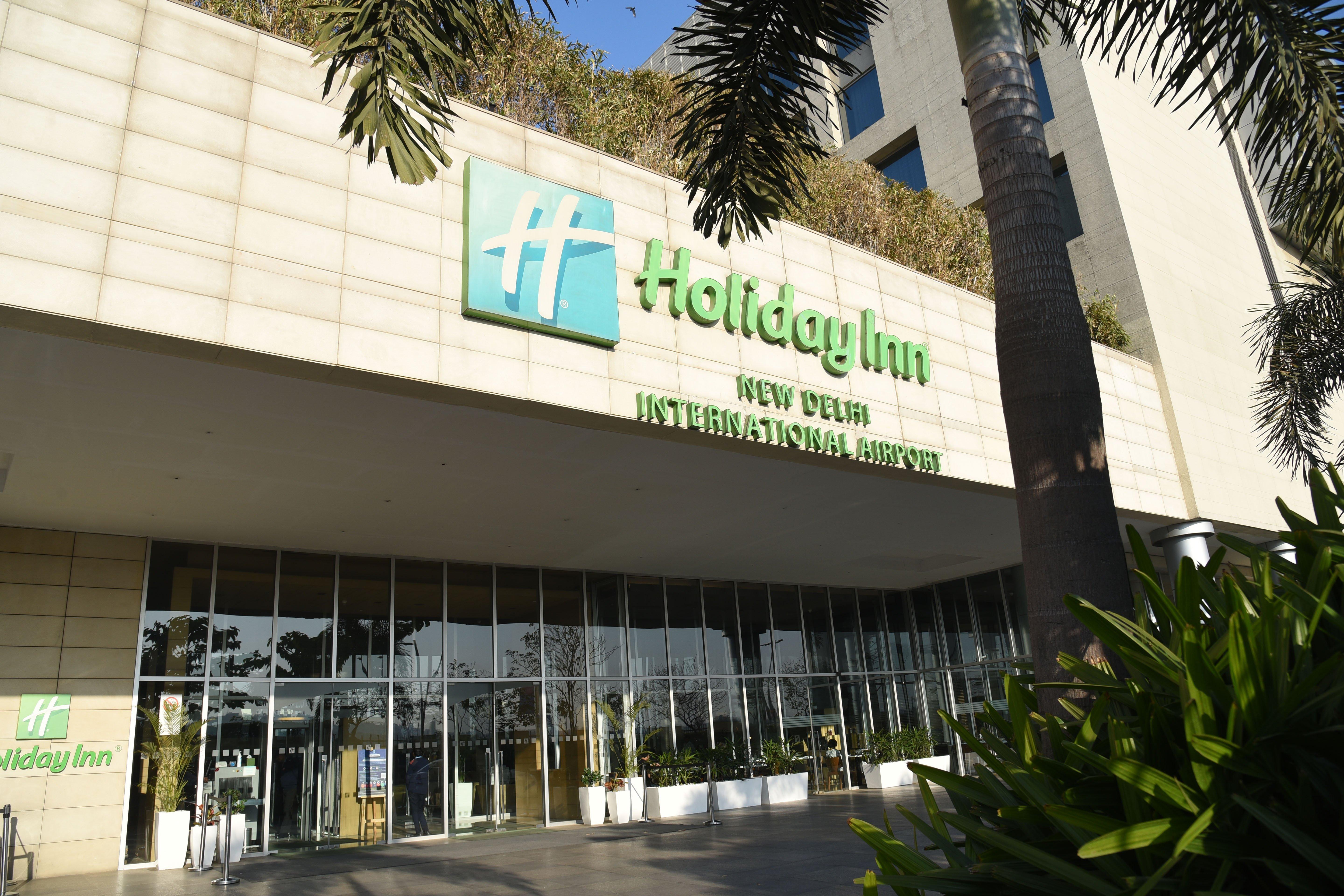 Holiday Inn New Delhi International Airport, An Ihg Hotel Exterior photo The photo shows the exterior of a Holiday Inn hotel, specifically the Holiday Inn New Delhi International Airport. The building features a modern design with a prominent green and white sign displaying the hotel name. There are large glass doors at t